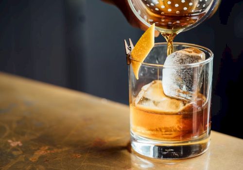 A cocktail being poured through a strainer into a glass with ice and an orange twist, set on a bar counter.