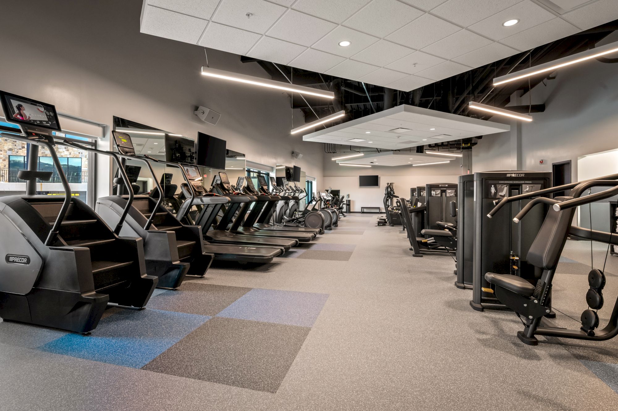 A spacious gym with rows of treadmills and weight machines, bright lighting, and a clean, modern layout. The ceiling has exposed tiles and workout equipment neatly organized.