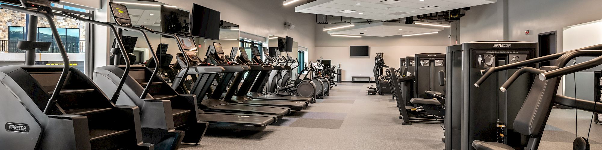A spacious gym with rows of treadmills and weight machines, bright lighting, and a clean, modern layout. The ceiling has exposed tiles and workout equipment neatly organized.