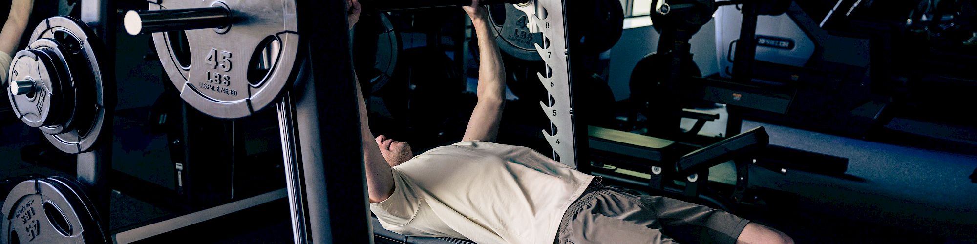 A person is bench-pressing on a gym bench with a barbell, plates on the sides, in a dimly lit workout room.