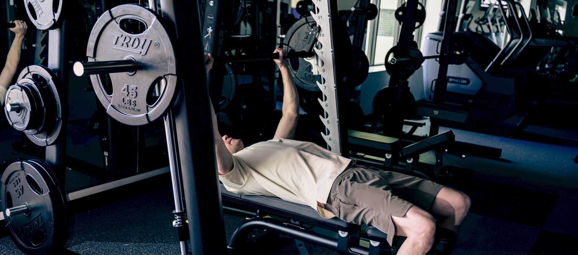 A person is bench-pressing on a gym bench with a barbell, plates on the sides, in a dimly lit workout room.
