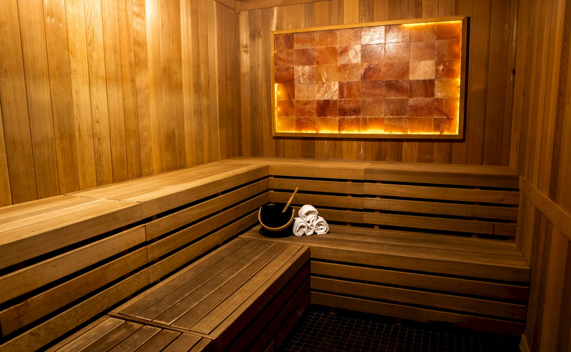 A wooden sauna room with tiered benches, a bucket, towels, and a lit salt brick panel on the wall ends the sentence.