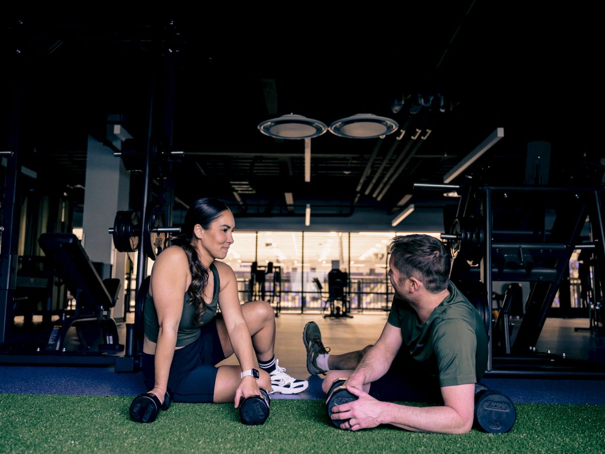 Two people sit on the gym floor, resting after a workout, talking and stretching near a mirrored workout space.