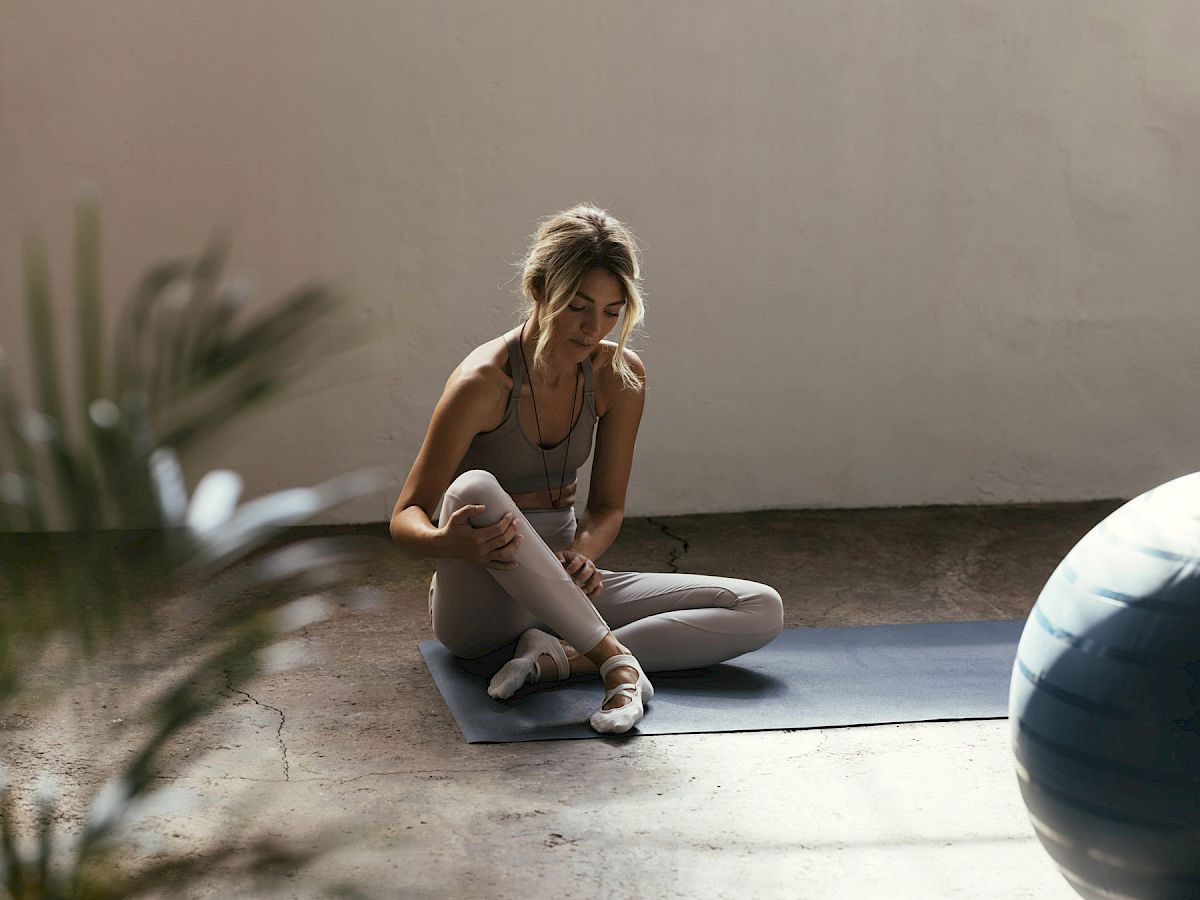 A person is sitting on a yoga mat in a sunlit room with a fitness ball nearby, surrounded by plants.