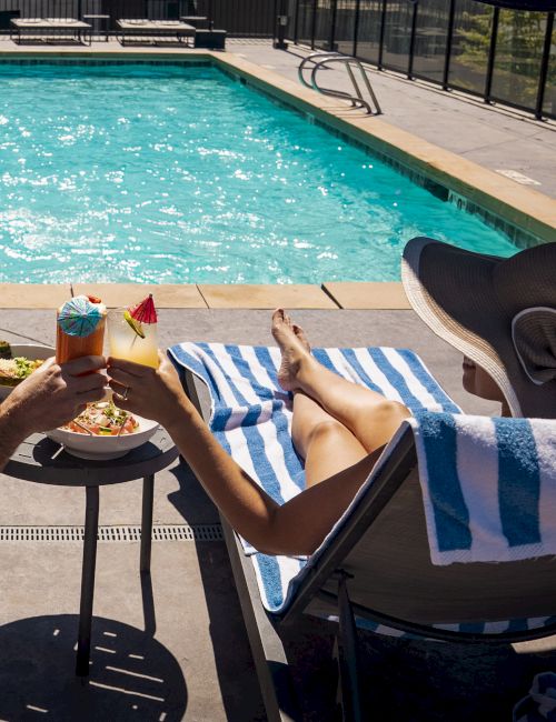 A poolside scene: someone lounging in a chair by the water, holding a tropical drink and a slice of pizza, with another drink on a side table, sunny and relaxing.