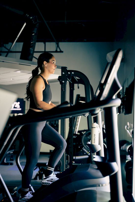 A woman runs on a treadmill in a gym, focused and mid-workout, with exercise machines and dim lighting in the background.