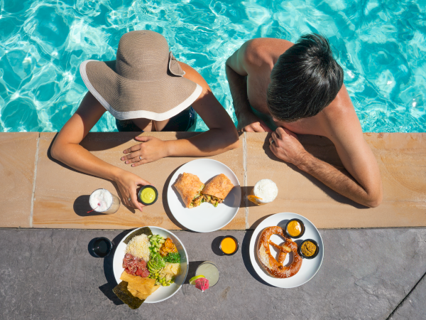 Two people dine poolside: a woman in a sunhat and a man lean over a table of tropical brunch with eggs, toast, salad, fruit, and juice.