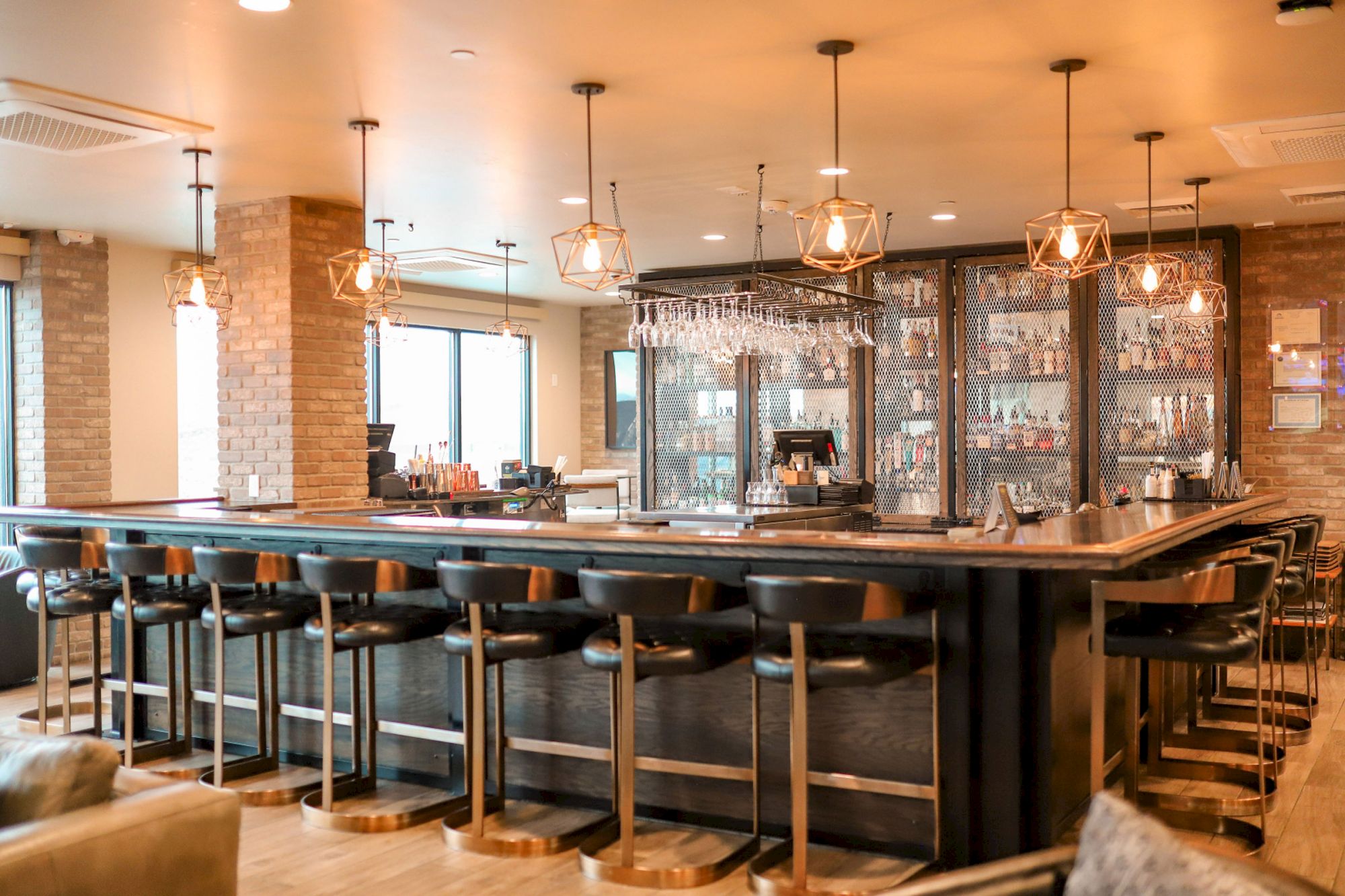 The image shows a modern bar with high chairs, hanging lights, and shelves stocked with glasses and bottles, creating a cozy atmosphere.