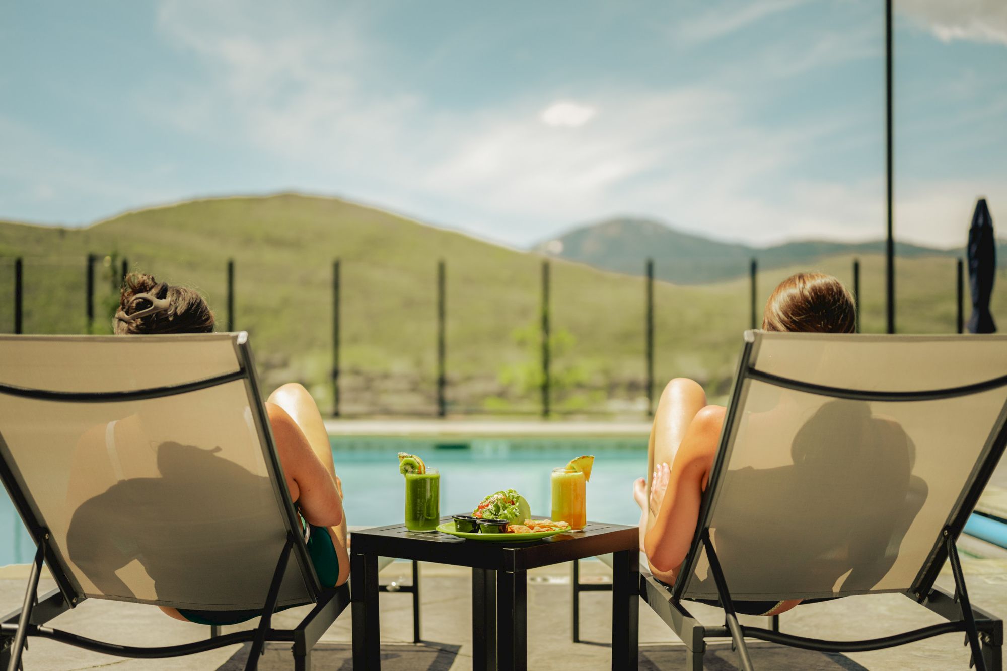 Relaxing by a pool, two people lounge in chairs with drinks and snacks, facing green hills and mountains under a cloudy sky.