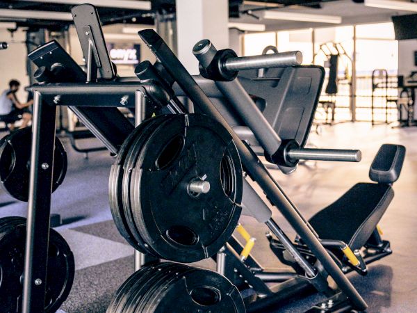 A gym machine rack with heavy plates loaded, a leg press/press combo, and a spacious workout area in the background.