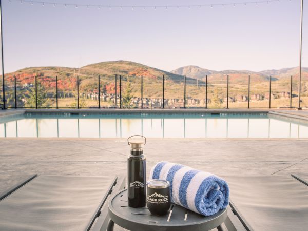 A pool with mountain views is in the background, and a table with a striped towel, a thermos, and a jar is in the foreground.
