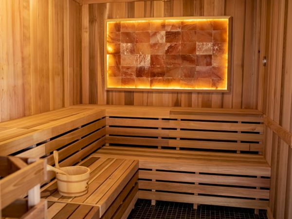 The image shows a wooden sauna room with benches, a bucket, a ladle, and an illuminated salt panel on the wall.