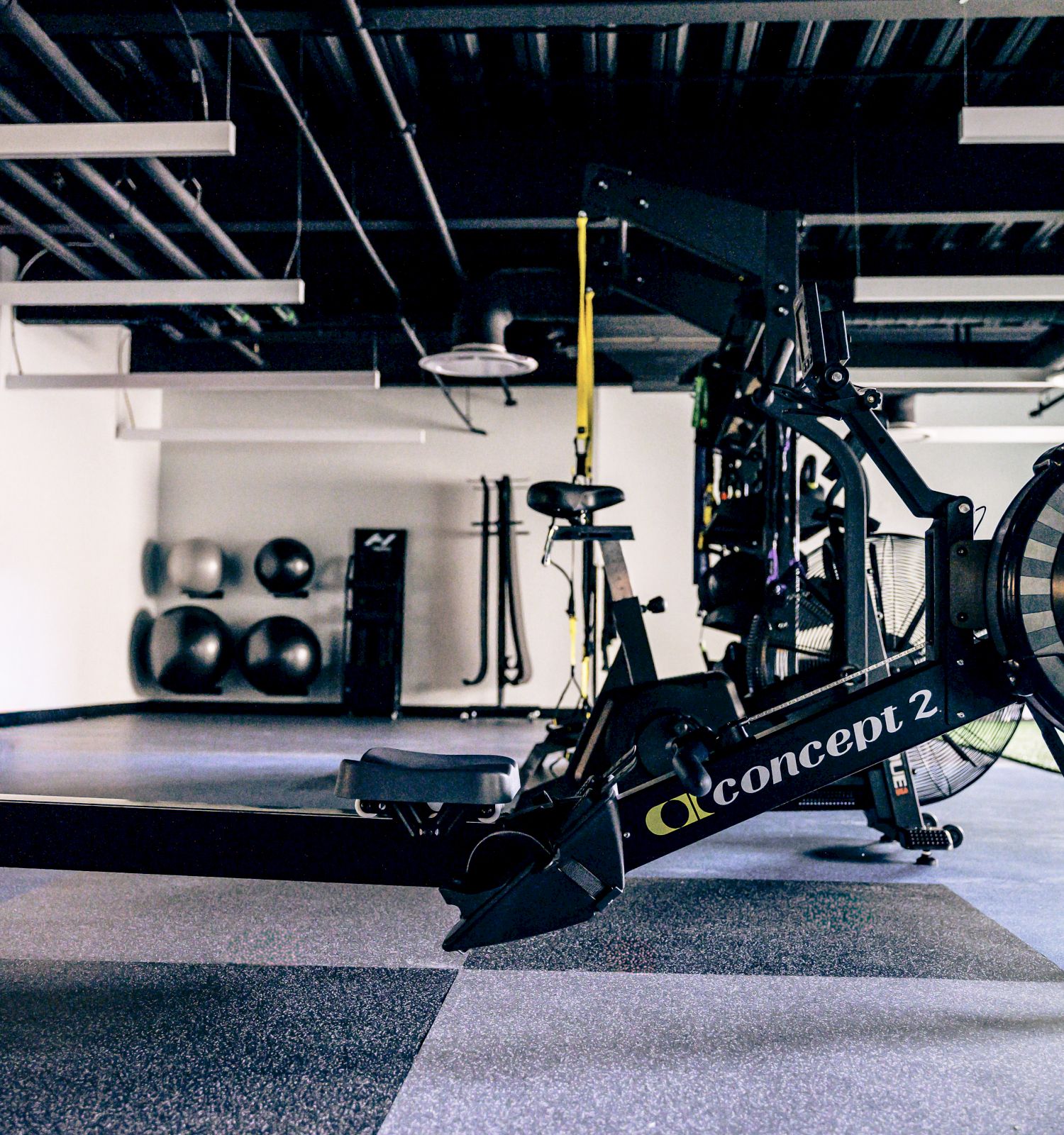 A Rowing Concept2 machine in a modern gym, with weights, resistance bands, and a fan in the background.