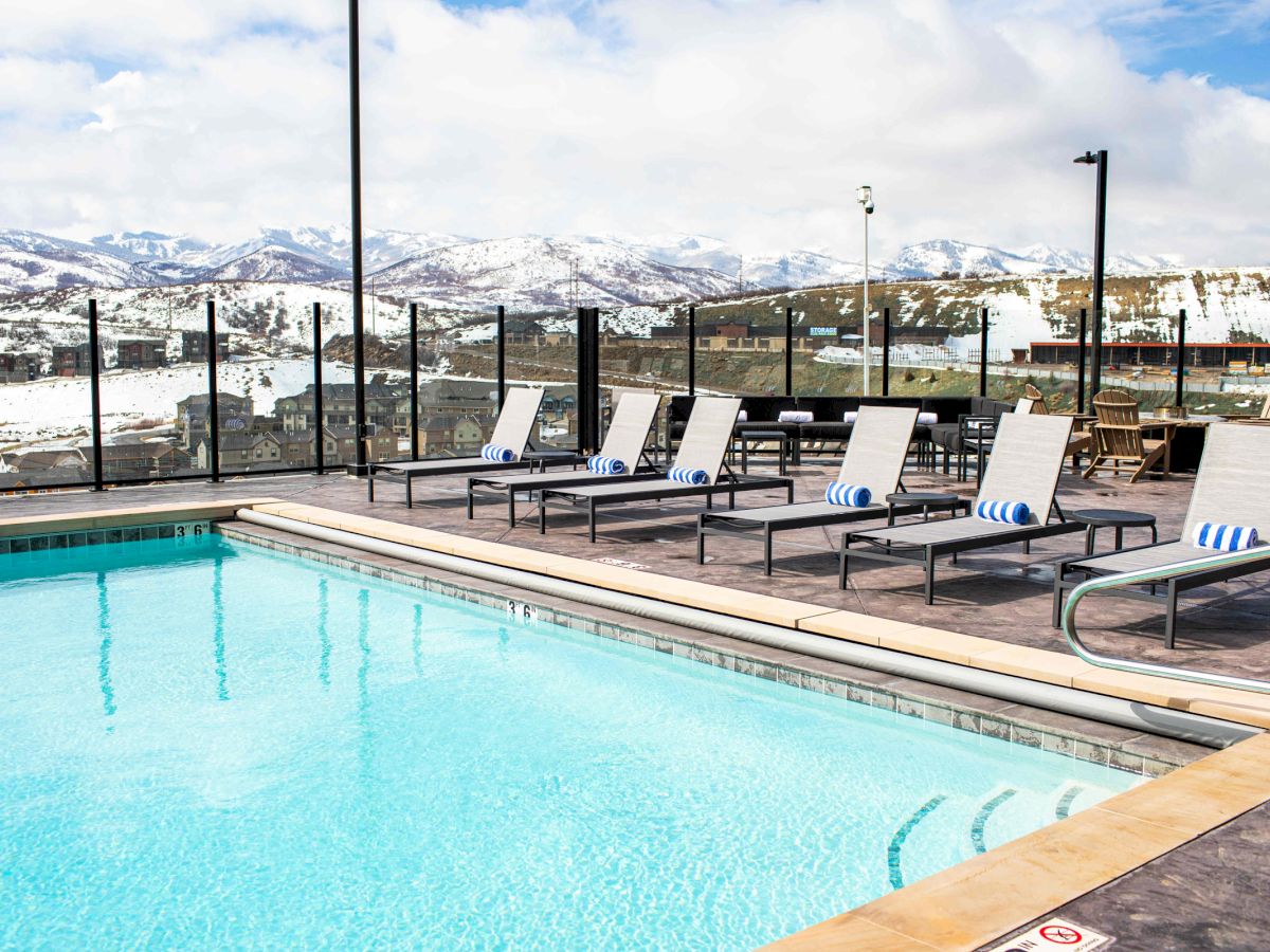 A pool with lounge chairs overlooks a scenic view of snow-covered mountains and a cloudy sky, surrounded by a glass fence.