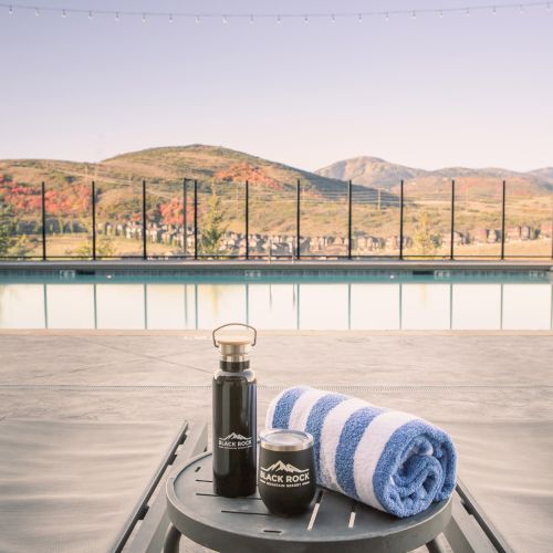 A poolside setting with a striped towel, thermos, and small jar on a table, against a backdrop of mountains and clear skies.