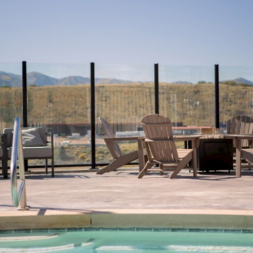 A poolside area with chairs and a table by a glass fence, overlooking hills under a clear sky.