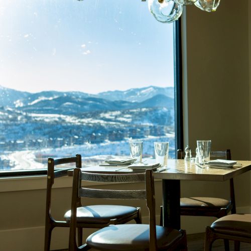 A dining area with a round table and chairs, set with glassware, and a large window showcasing a mountain view under a unique chandelier.