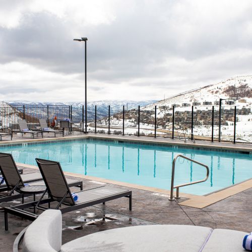 A poolside area with lounge chairs overlooks snowy mountains under a cloudy sky.