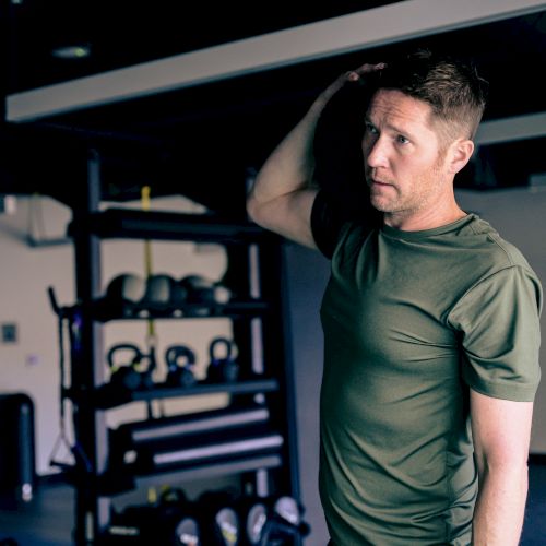 A man in a green shirt stands in a dim gym with weights and equipment behind him, looking upward.