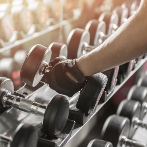 A person wearing a glove is picking up a dumbbell from a rack in a gym setting, with a mirror reflecting the equipment.