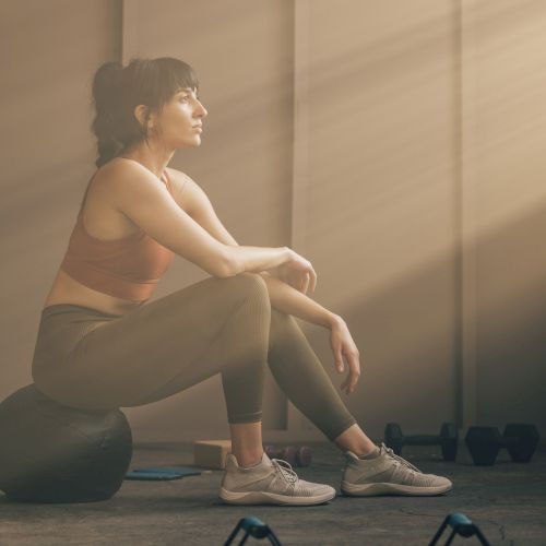 A person in workout attire rests on a gym ball, surrounded by dumbbells, under sunlight streaming through a window.