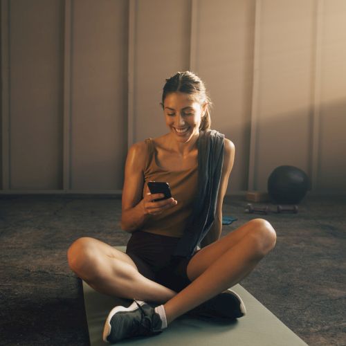 A person sits cross-legged on a yoga mat, smiling at their phone, with a towel over their shoulder, in a softly lit space.