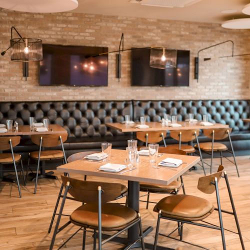 The image shows a modern restaurant interior with a brick wall, cushioned bench seating, wooden tables, chairs, and industrial-style lights.