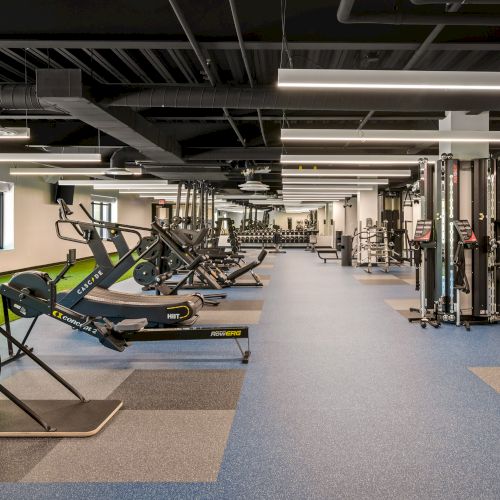 A gym with people working out, weight machines, and bright lighting, plus open space and mirrors at the back.