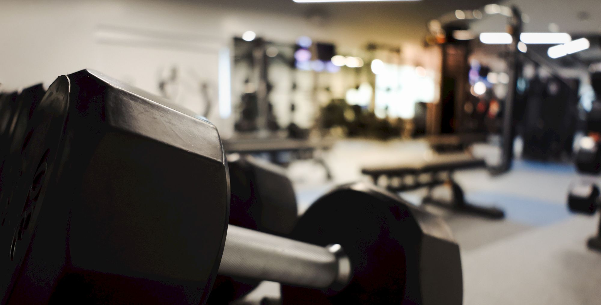 A close-up of a dumbbell in a gym foreground with blurred workout equipment and benches in the background.