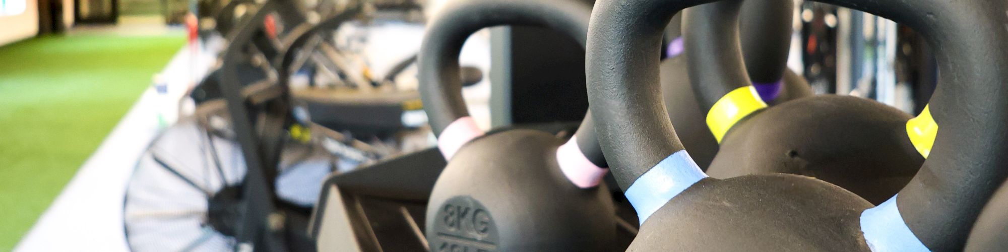 Kettlebells labeled 12 KG sit on a rack in a modern gym, with fans and exercise equipment in the background under bright ceiling lights.