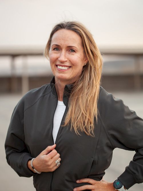 A smiling woman with long blond hair wearing a black jacket, standing confidently with one hand on her hip, outdoors on a sunny day.