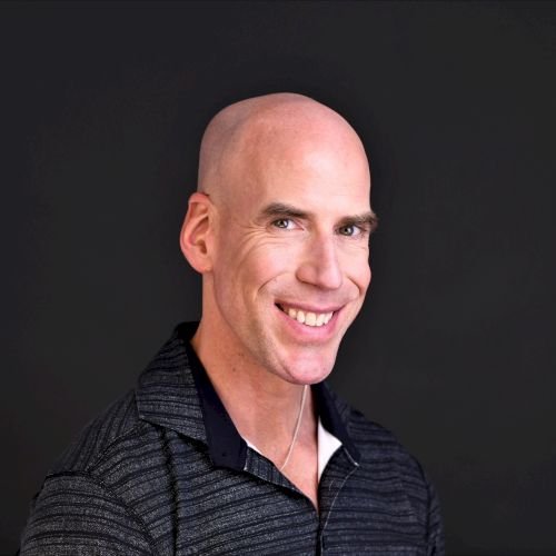 A smiling bald man in a dark, collared polo shirt posing against a dark background.