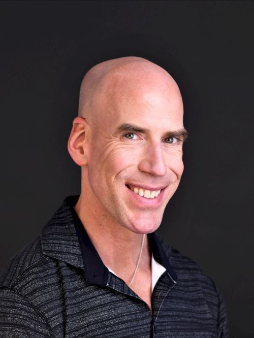 A smiling bald man in a dark, collared polo shirt posing against a dark background.
