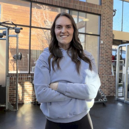 A young woman with long dark hair stands in a sunlit, brick-walled entryway wearing a light blue hoodie and black leggings, smiling with arms crossed.