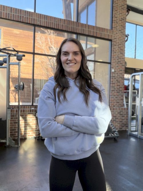 A young woman with long dark hair stands in a sunlit, brick-walled entryway wearing a light blue hoodie and black leggings, smiling with arms crossed.