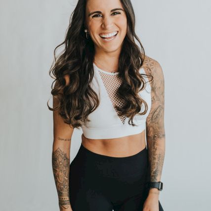 A smiling woman with long dark wavy hair, wearing a white crop top and black mesh leggings, standing against a light background.