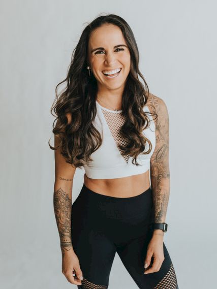 A smiling woman with long dark wavy hair, wearing a white crop top and black mesh leggings, standing against a light background.