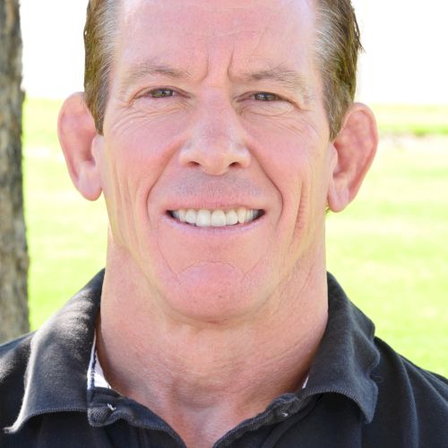 A smiling man with short brown hair wearing a dark polo, outdoors in a sunny park setting, neutral expression, erect posture.