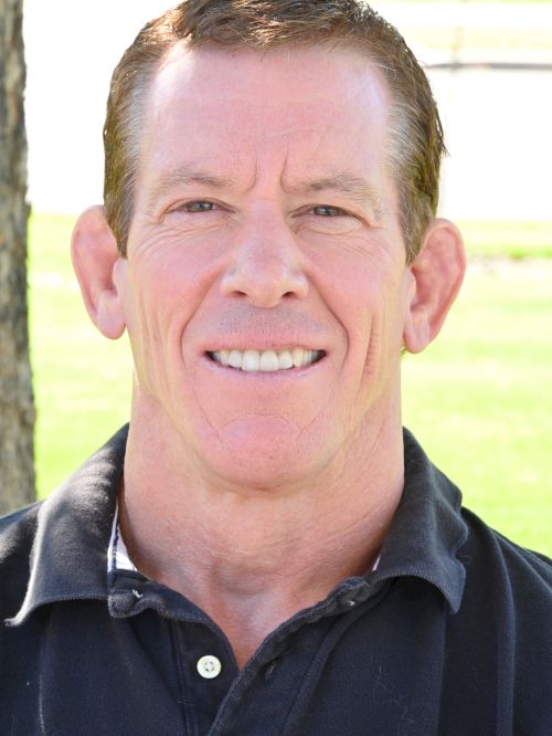 A smiling man with short brown hair wearing a dark polo, outdoors in a sunny park setting, neutral expression, erect posture.