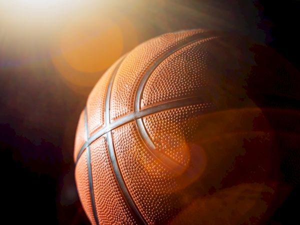 A close-up of a basketball with textured orange surface and black seams, lit dramatically from the side.
