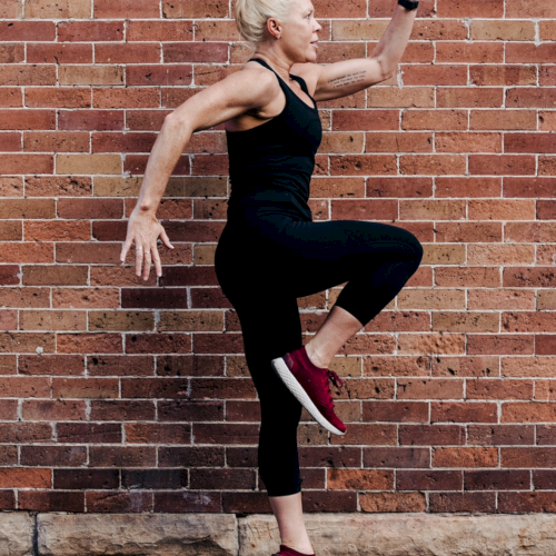 A fit woman in black athletic wear leaps with one knee raised, arms poised, against a brick wall near a sidewalk.