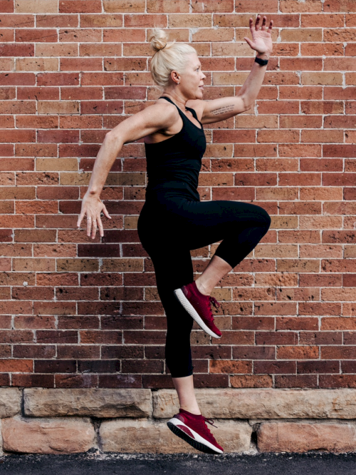 A fit woman in black athletic wear leaps with one knee raised, arms poised, against a brick wall near a sidewalk.