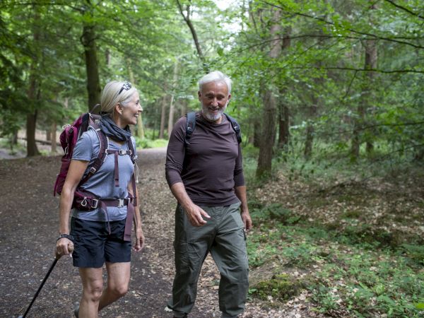 Two hikers, a smiling woman with a trekking pole and a bearded man, walk along a wooded trail, backpacks on, enjoying the outdoors.