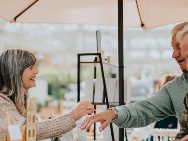 Two people smiling at a market or shop, exchanging a handshake or payment near a display under an umbrella, with a warm, friendly vibe.