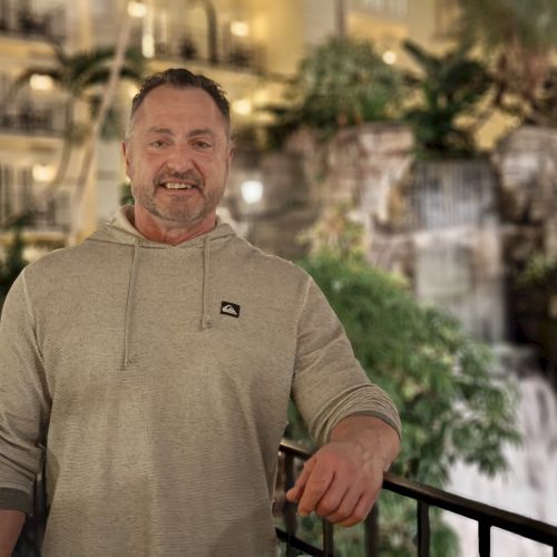 A smiling man in a light gray hoodie stands on a railing indoors, with greenery and a fountain-like backdrop behind him.