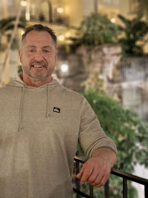 A smiling man in a light gray hoodie stands on a railing indoors, with greenery and a fountain-like backdrop behind him.