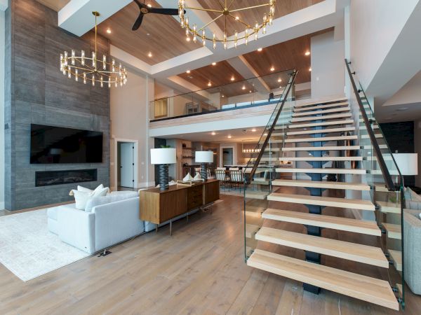 A spacious modern living area with a floating wooden staircase, steel railing, high ceilings, and a two-story open plan accented by pendant chandeliers.