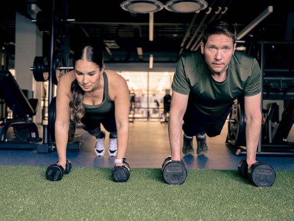 A man and woman in a gym hold a plank position on dumbbells, working out on artificial turf in the background.