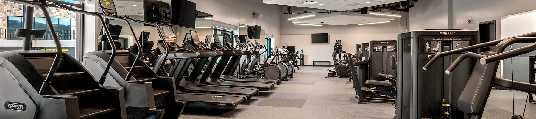 A modern gym with rows of cardio machines—treadmills and ellipticals—plus strength stations under bright LED lights.
