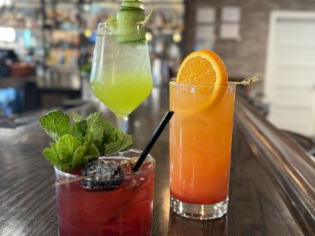 Three colorful cocktails sit on a wooden bar: green, orange with an orange slice garnish, and red with mint and berries.